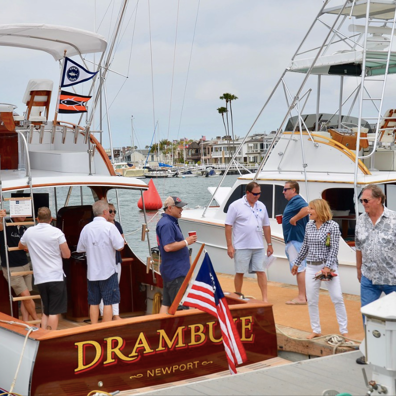 People socializing on a yacht docked in a marina, with “DRAMBUIE” on the stern and sailboats nearby.