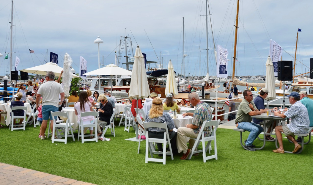 Outdoor seating area with people at white tables on a grassy lawn under a cloudy sky, with boats in the background