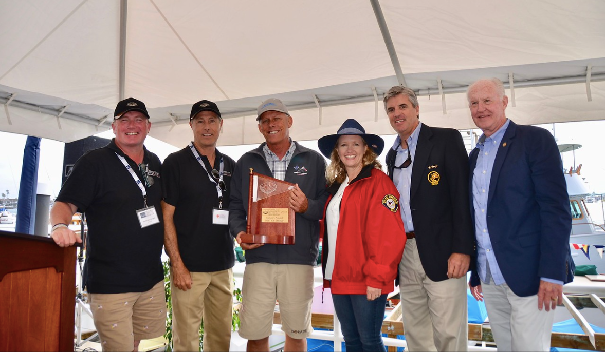 Six people on a stage hold an award plaque under a white tent, smiling at a ceremony