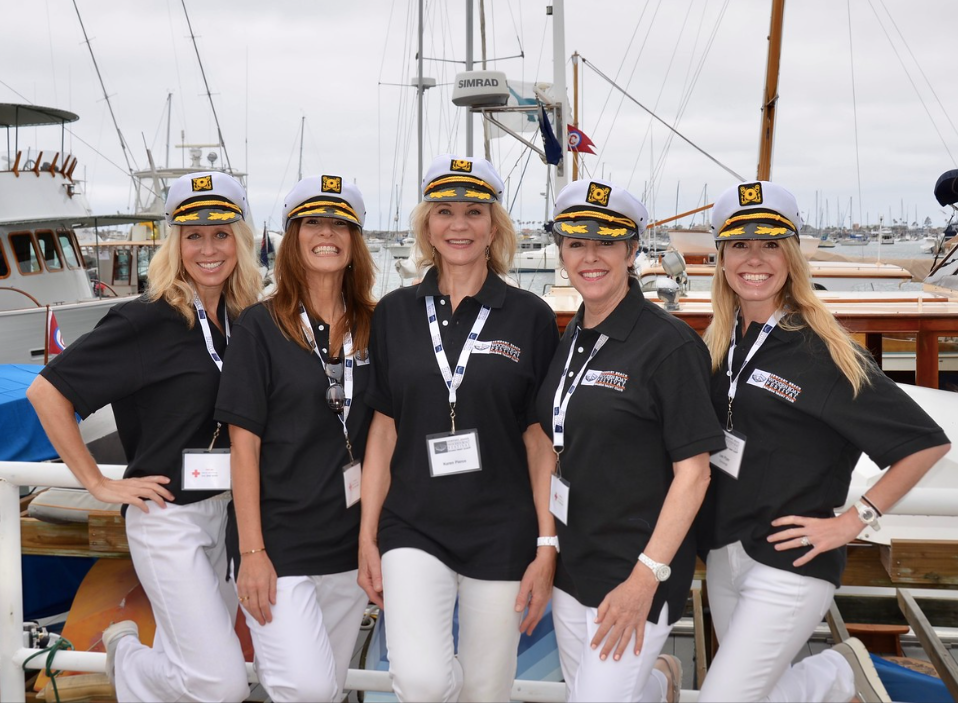 Five women in black shirts and white pants smiling on a docked boat, with masts and sailboats behind them