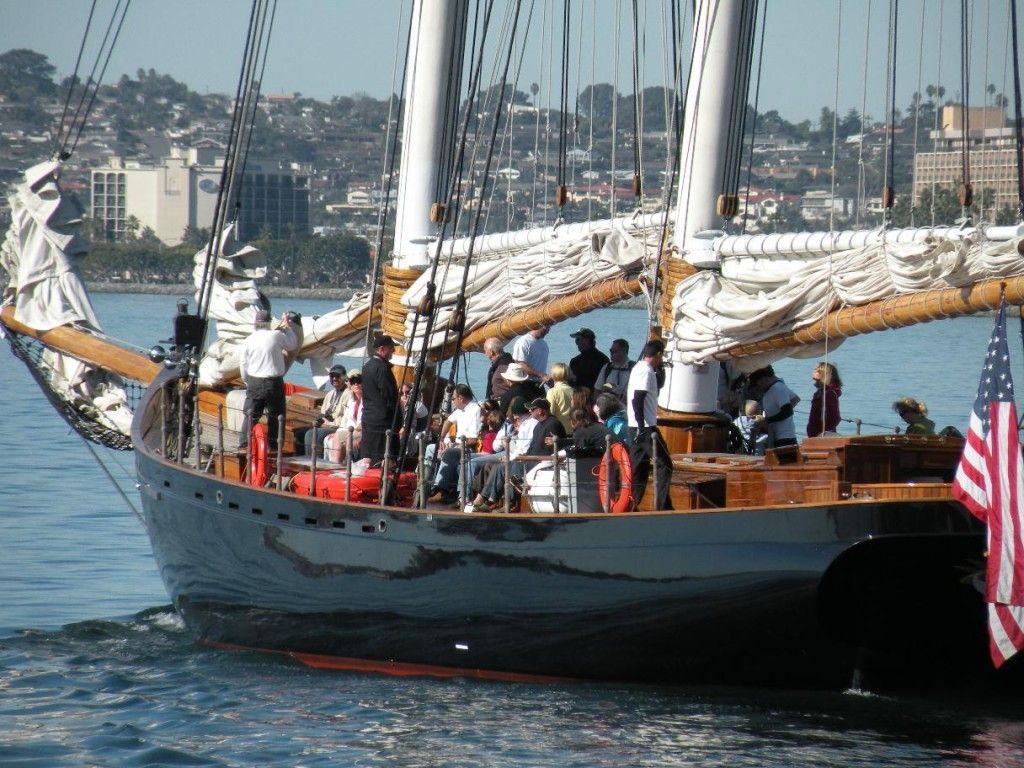 Large wooden sailboat with people on deck on calm water, city skyline in the background