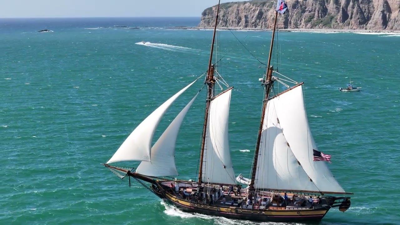 Two-masted sailboat with white sails on turquoise water near rocky coastline. Spirit of Dana Point Boat.