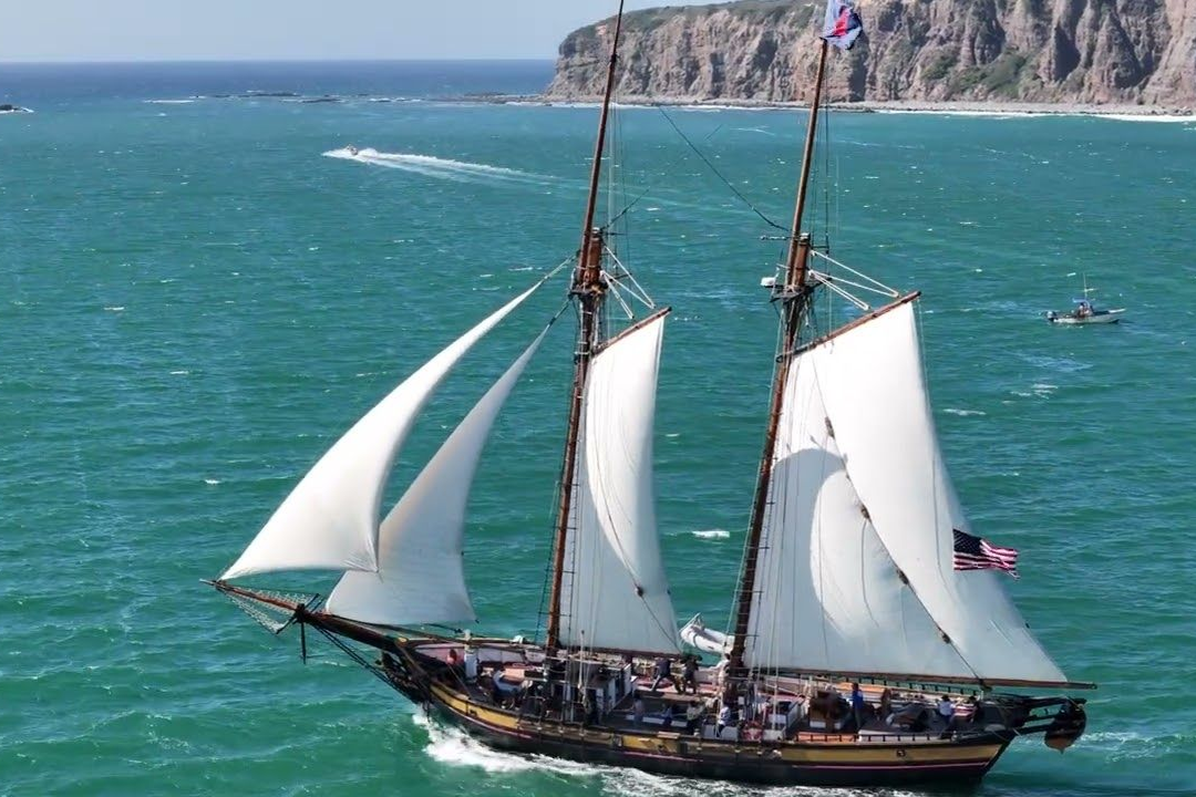 The Spirit of Dana Point, tall sailboat with white sails on turquoise sea near a rocky coastline