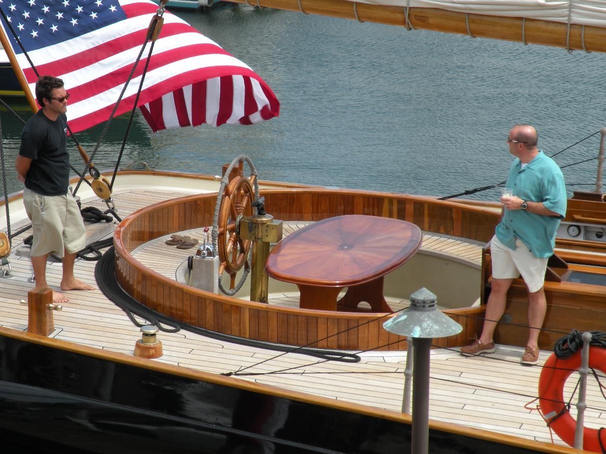 Two people on a sailboat deck with a large U.S. flag, steering wheel, and calm water backdrop