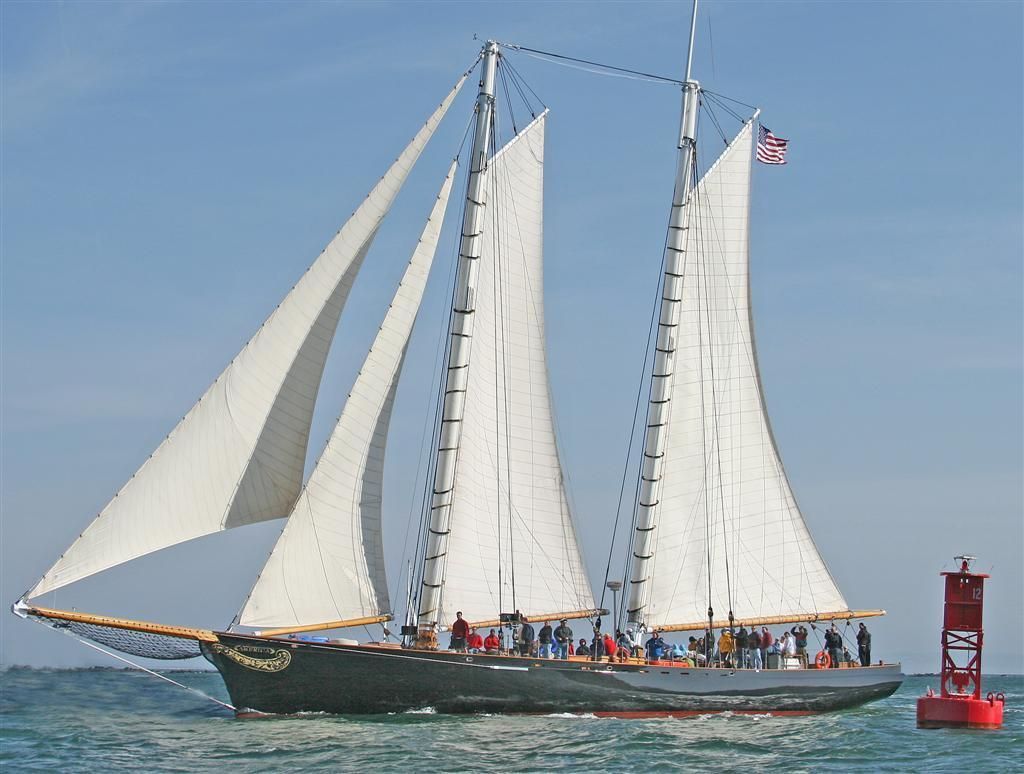 Large white-sailed boat with people aboard cruising on blue water near a red buoy