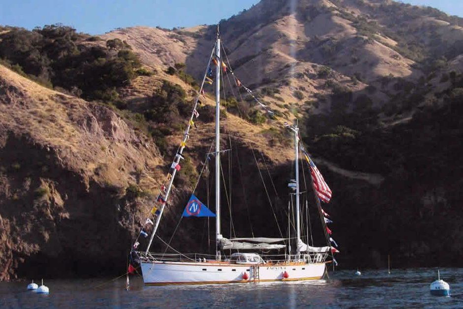 Sailboat anchored in a rocky cove beneath steep hills, with flags and calm water.