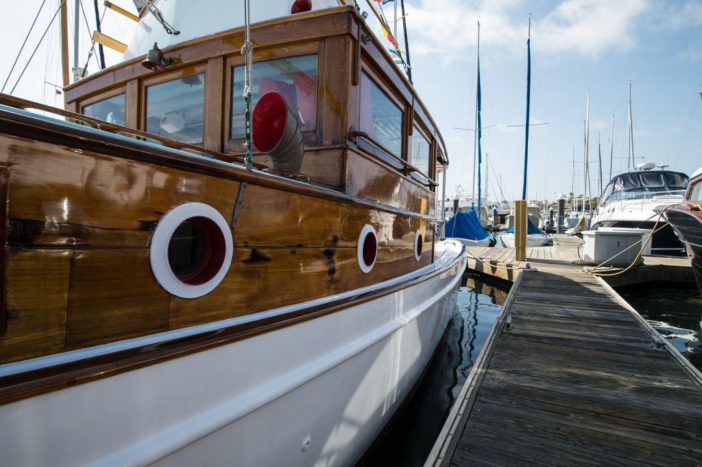 Wooden sailboat moored at a marina beside a dock, with yachts in the background under a clear sky.