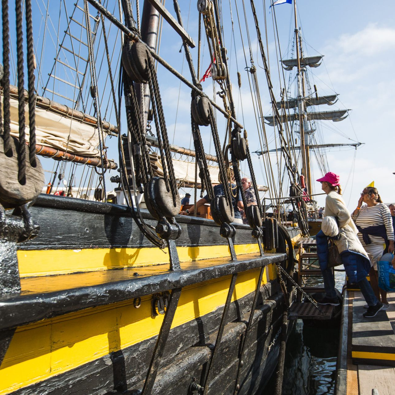 Sailors on a tall ship deck with black rigging and yellow hull, under a clear blue sky