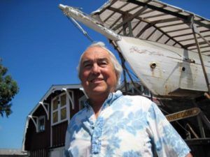 Man smiling outdoors beside a white sailboat under a blue sky