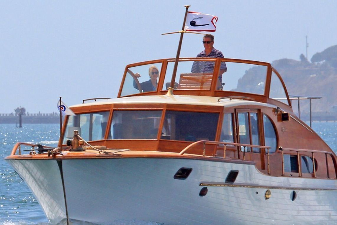 Wooden motor yacht on calm water with a person standing on deck and a flag flying above