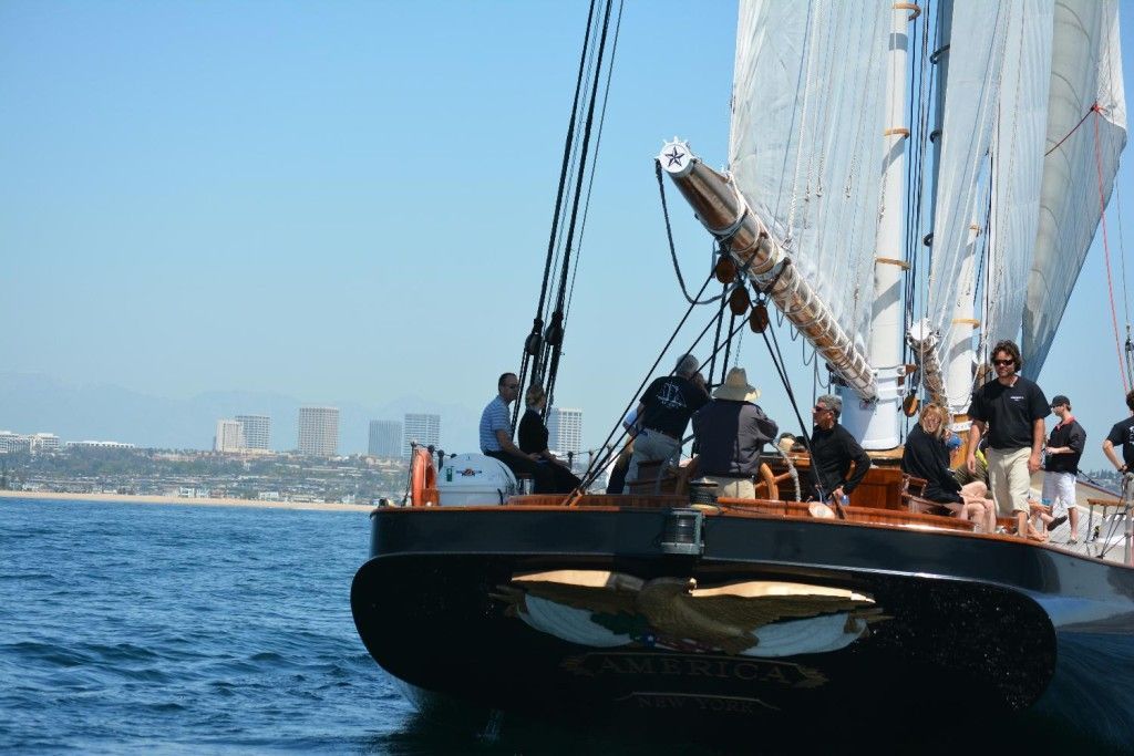 Sailboat with crew on blue water near a city skyline under a clear sky