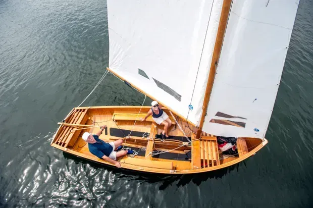 Two people sailing a small wooden boat on calm water, viewed from above.