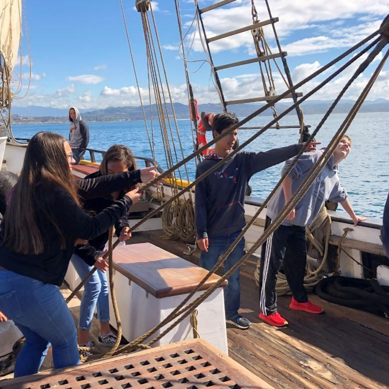 People handling ropes on a sailboat deck with water and sky in the background