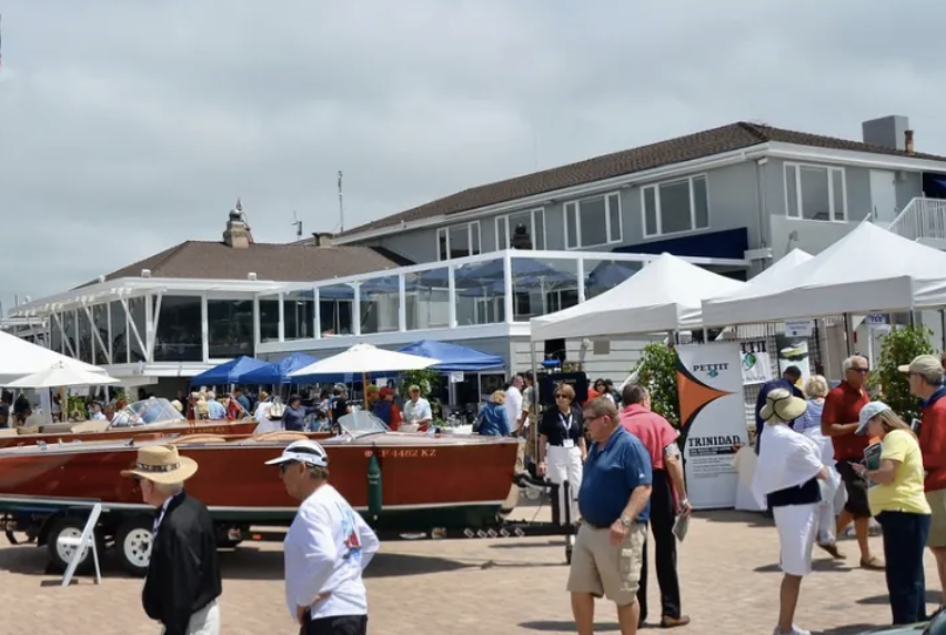 Crowded seaside marina with people, white tents, and boats in front of a waterfront building