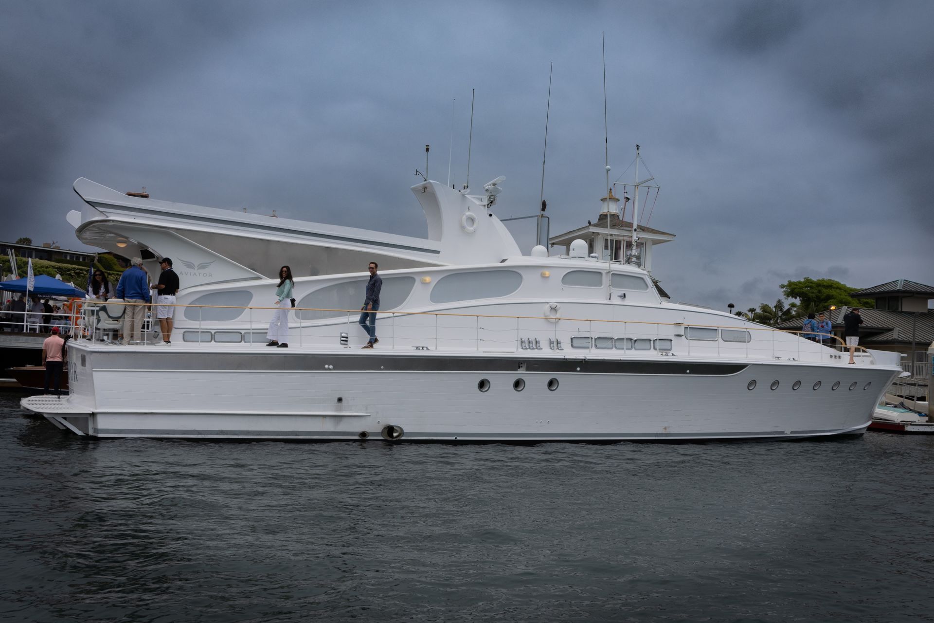 Large white yacht docked at a marina under a cloudy sky, with people on the deck and shore.