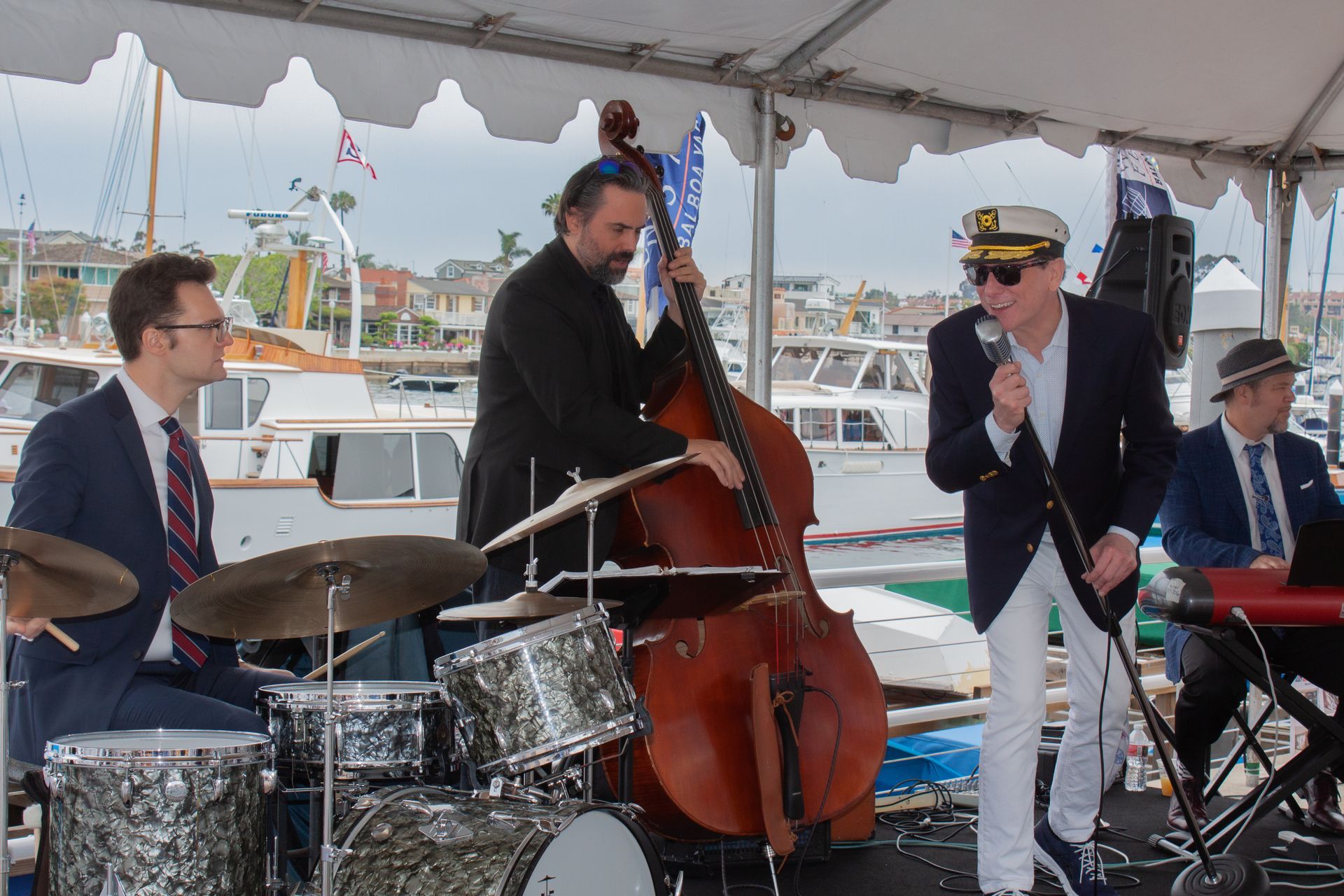 Jazz band performing under a white canopy by the waterfront, with drums, upright bass, keyboard, and singer.