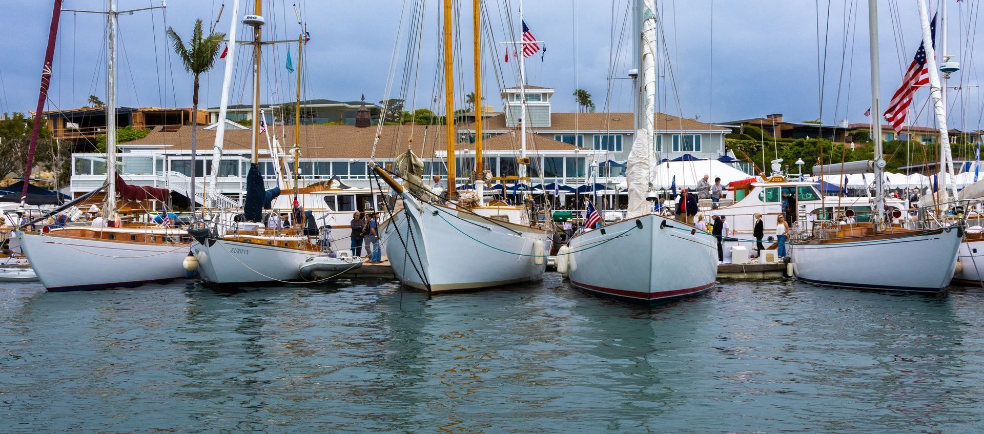 Sailboats docked at a marina with masts, calm water, and a cloudy sky.