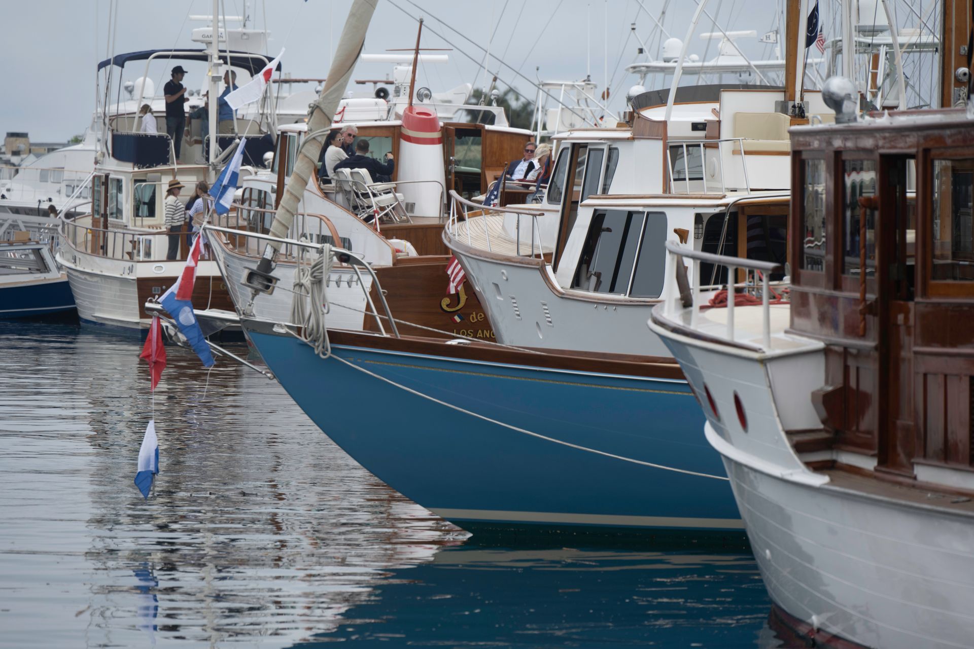 Docked boats at a harbor, including a blue sailboat and a wooden yacht, on calm water.