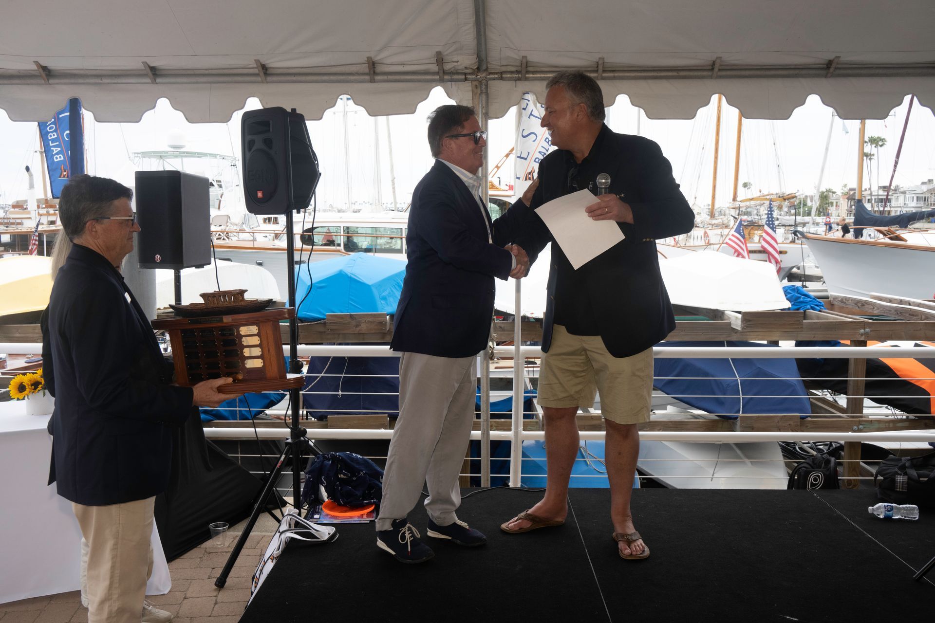 Two men shaking hands on a dockside stage, with a woman standing nearby and boats in the background.