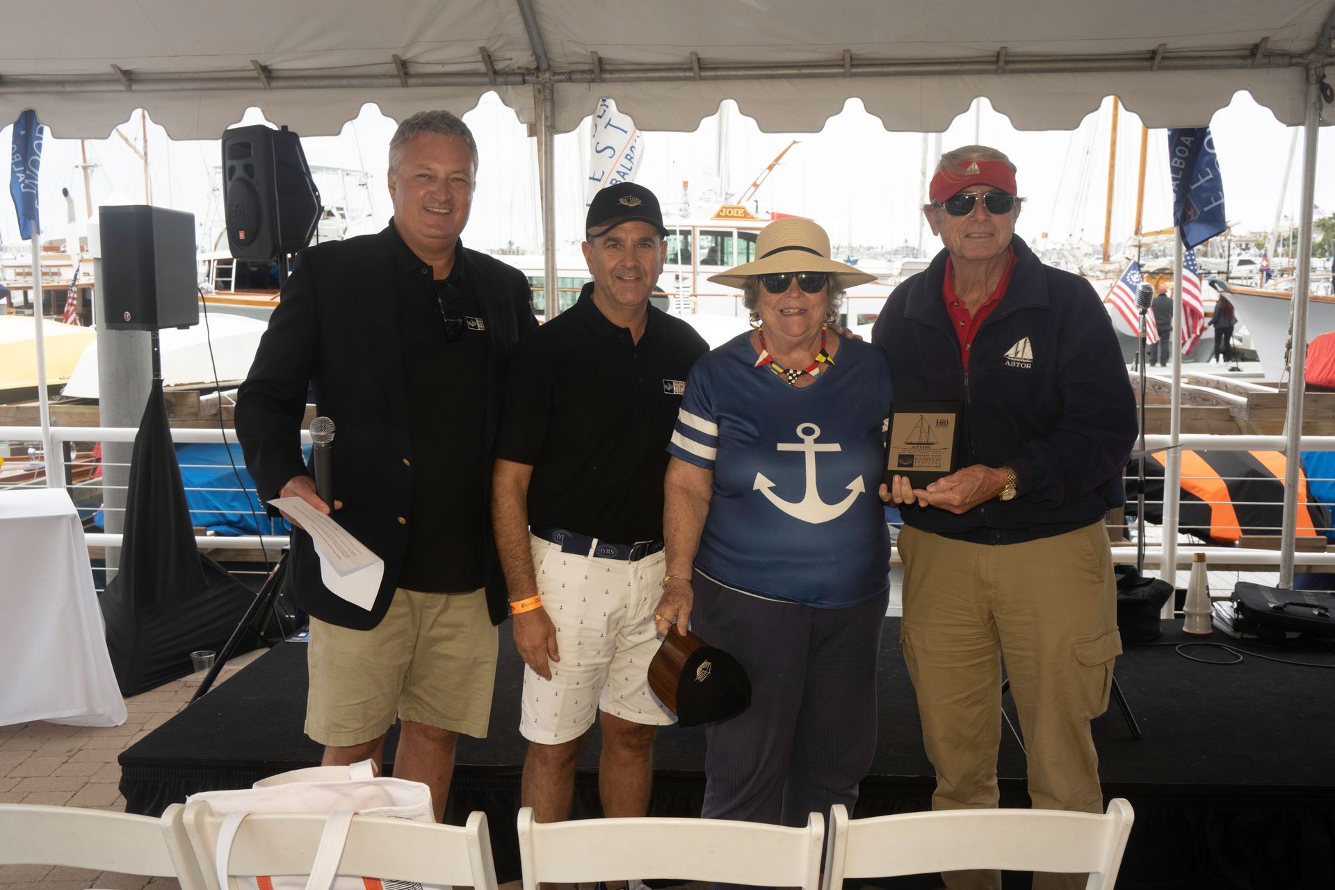 Four people posing under a tent at a marina event, with boats and tables in the background.