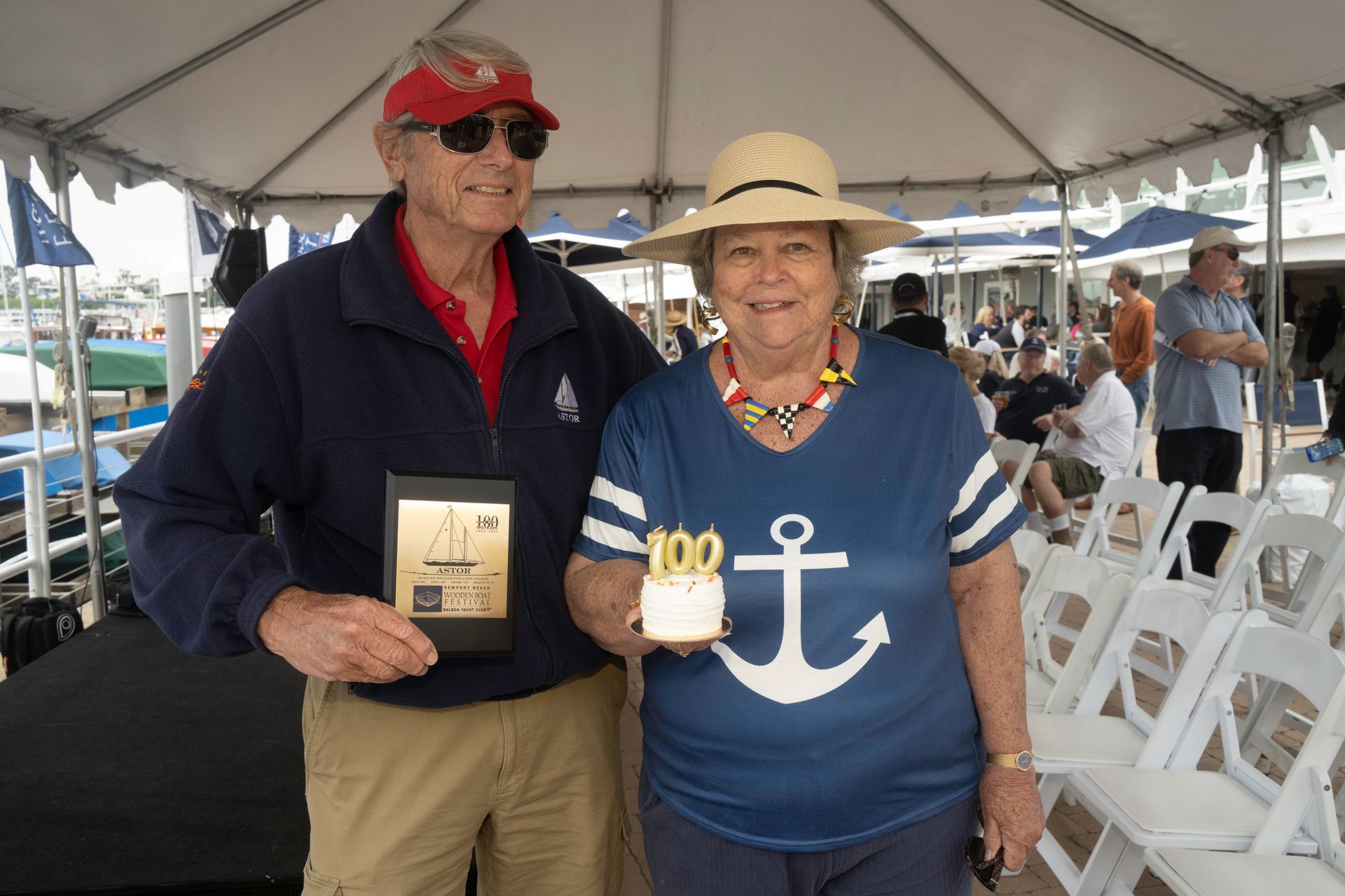 Two people standing under a tent, smiling and holding a plaque and mug, with rows of white chairs behind them.