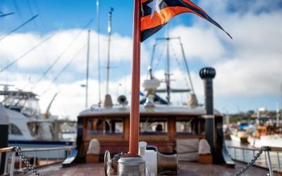 Docked wooden boat with a mast and black-orange flag, marina yachts in the background under a cloudy blue sky