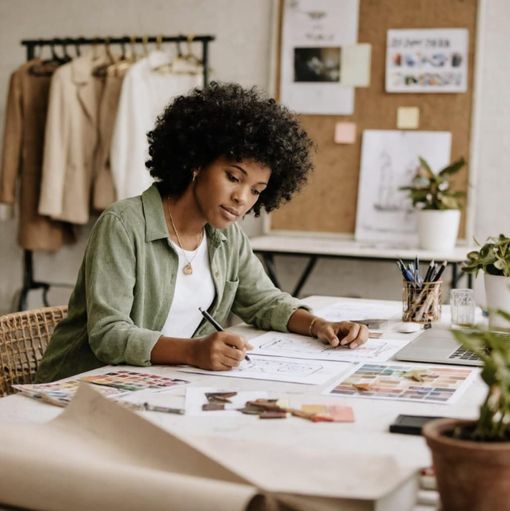 Person sketching fashion ideas at a desk in a bright studio with fabrics, mood boards, and plants