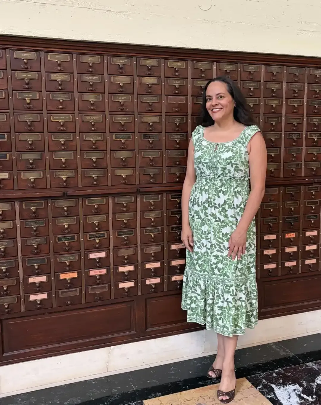 Woman in a green floral dress standing beside a wall of mailboxes, smiling indoors.