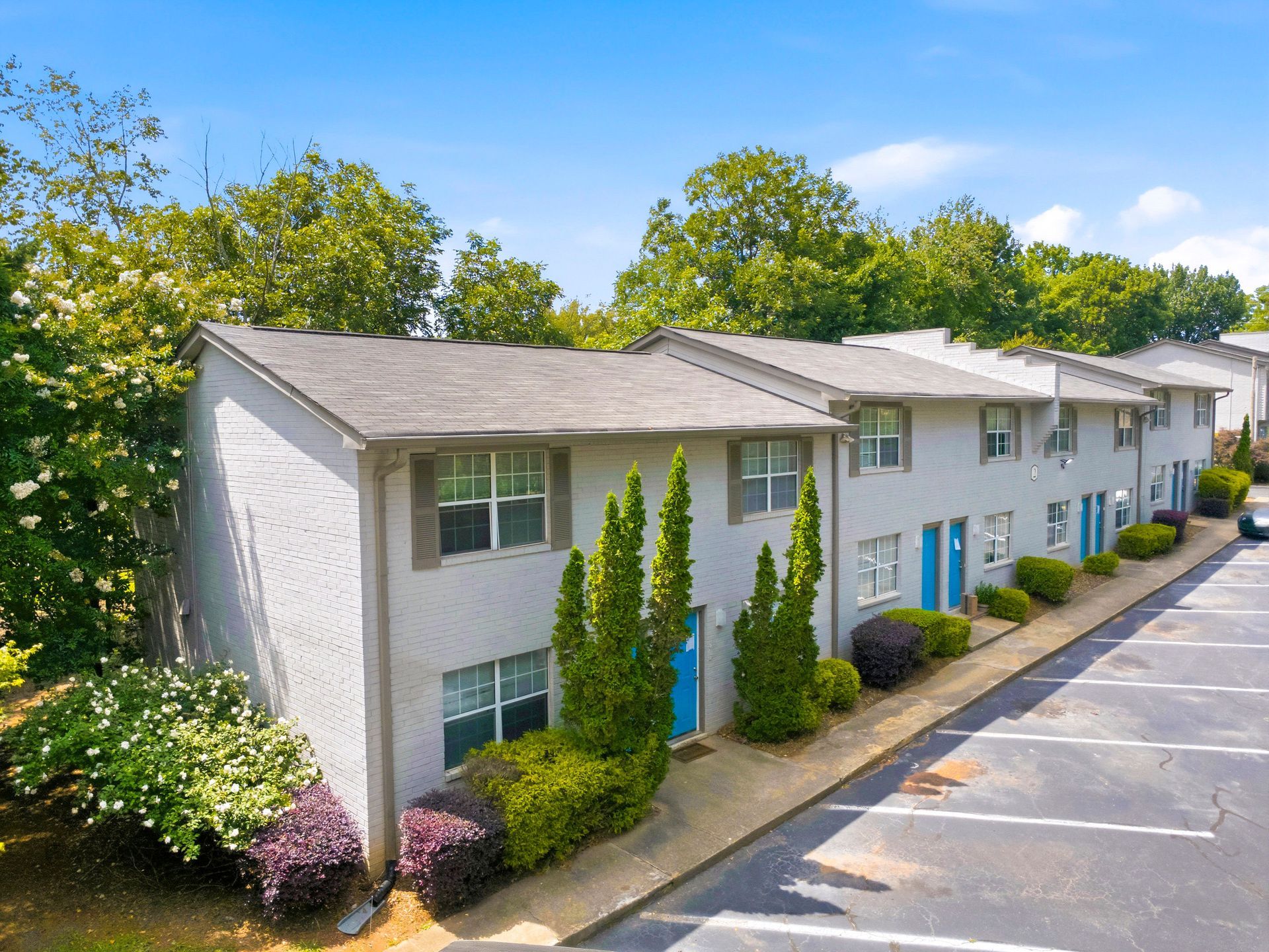 Apartment building with white siding, blue doors, and parking lot beside green trees