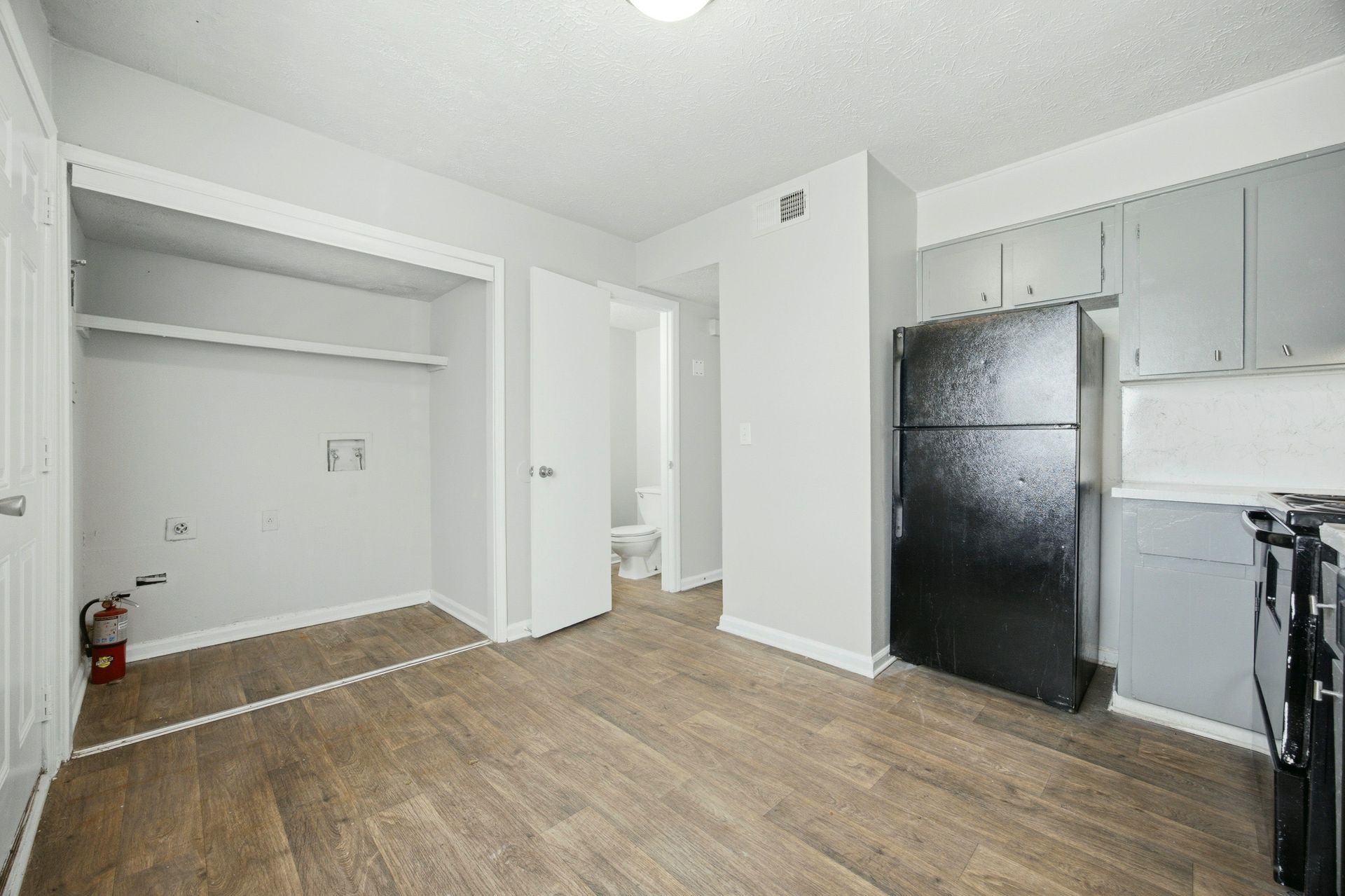 Empty apartment kitchen and living area with wood floors, white walls, and a black refrigerator