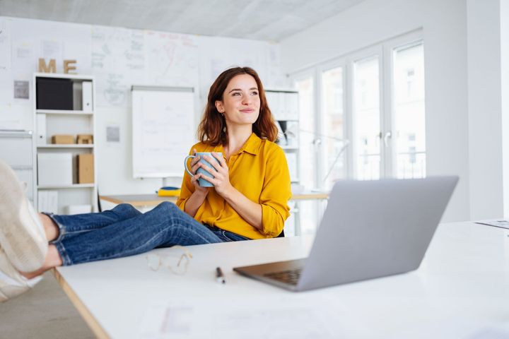 Woman in a bright home office with a laptop, holding a mug and reclining on a desk