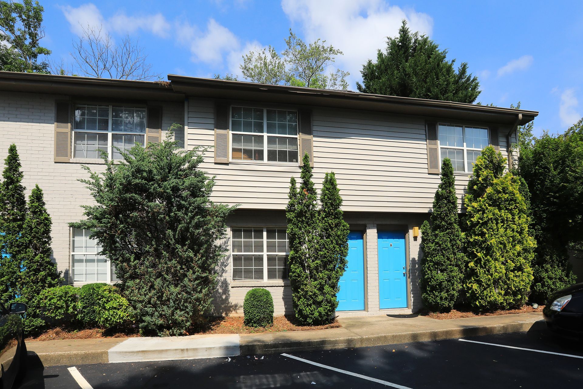Two-story beige townhouse with blue front doors, trees, and a parking lot under a blue sky