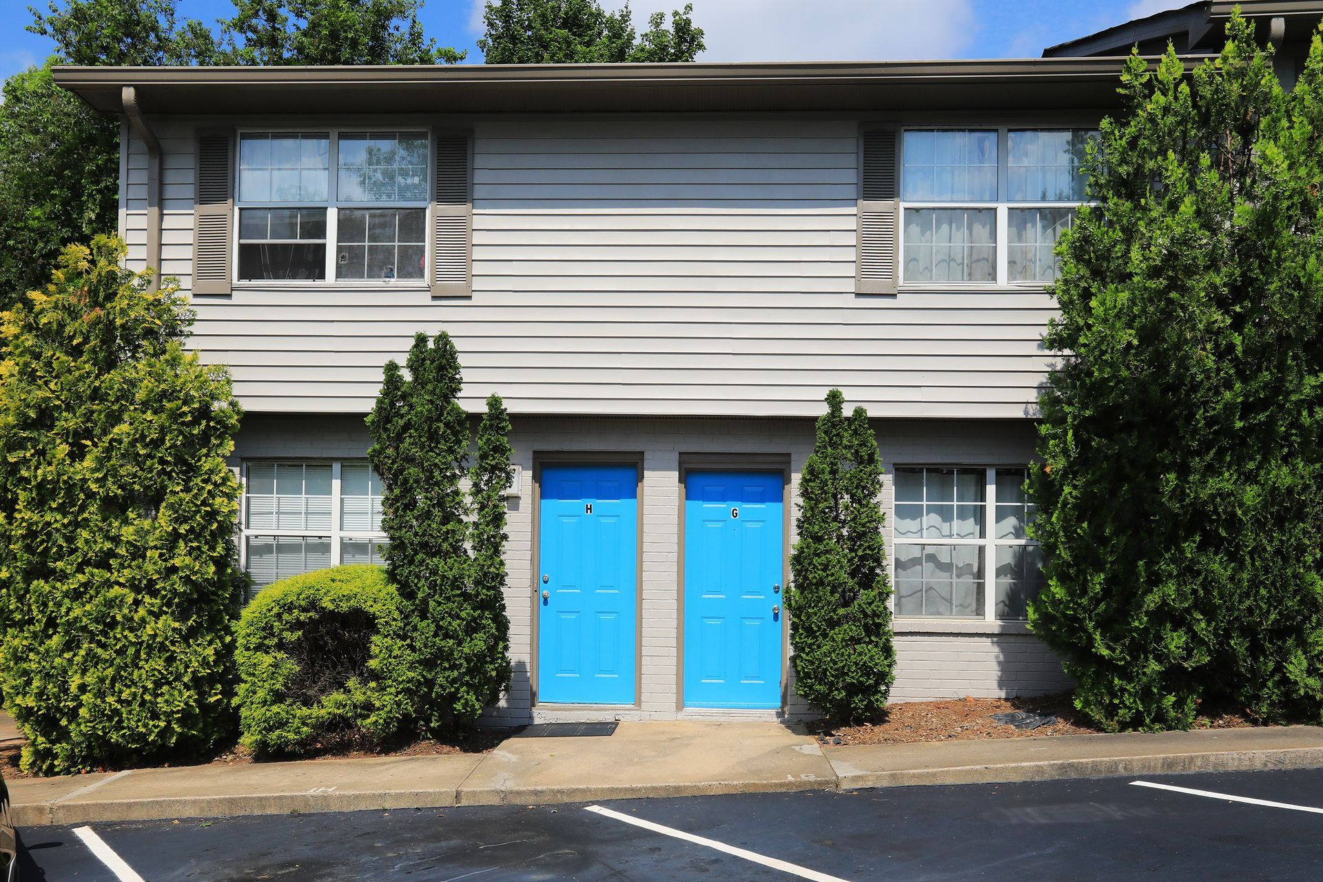 Two-story apartment building with bright blue double doors, surrounded by shrubs and trees.
