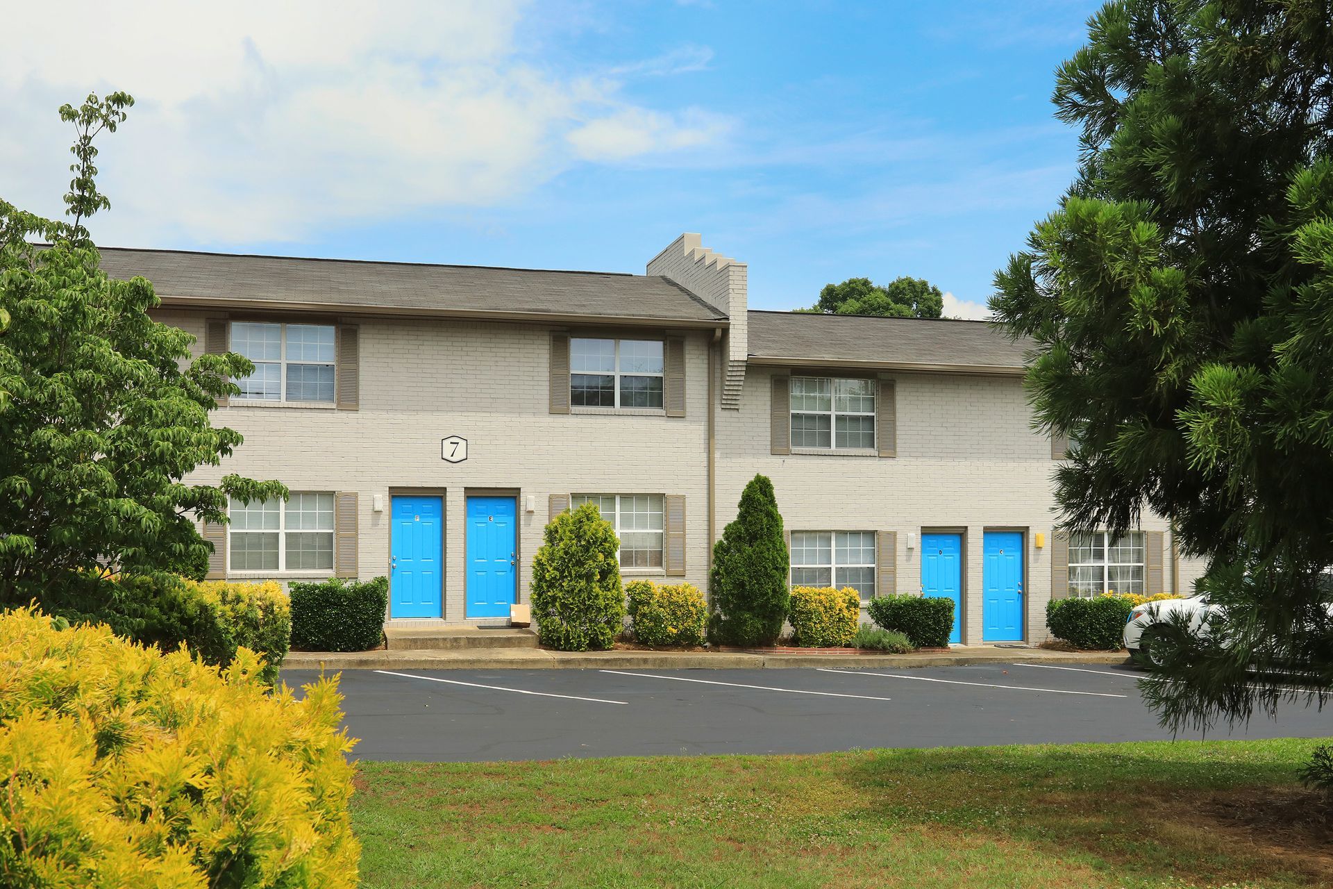 Two-story apartment building with blue doors and windows, surrounded by trees and shrubs