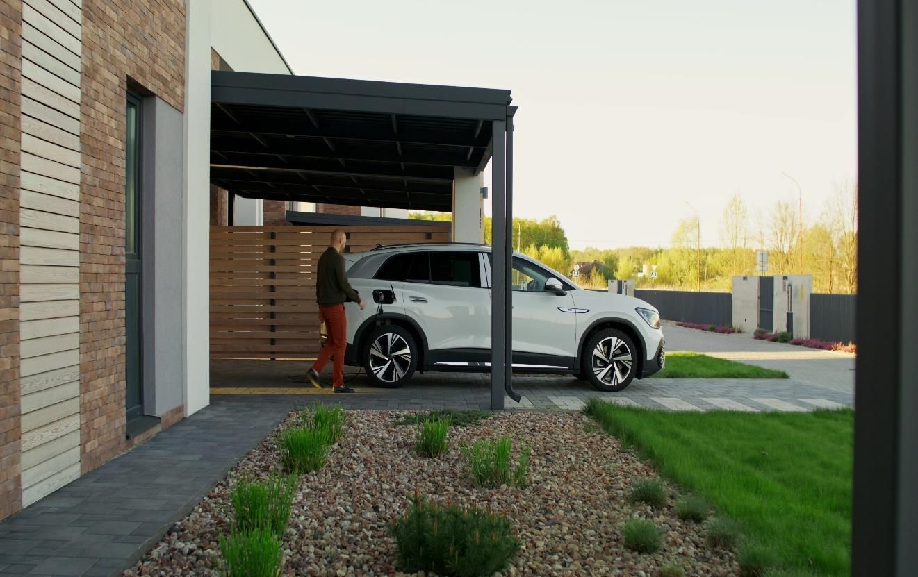White SUV parked under a carport beside a brick house, with a person standing nearby.