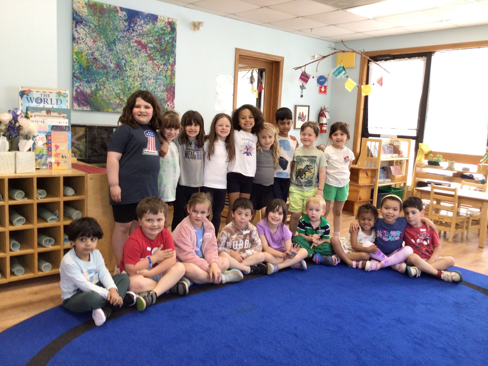 Group of children posing in a classroom with a blue carpet, colorful decorations, and shelves in the background.