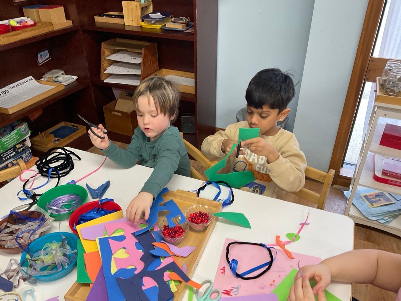 Children making crafts at a table with colorful paper, scissors, and supplies in a classroom.