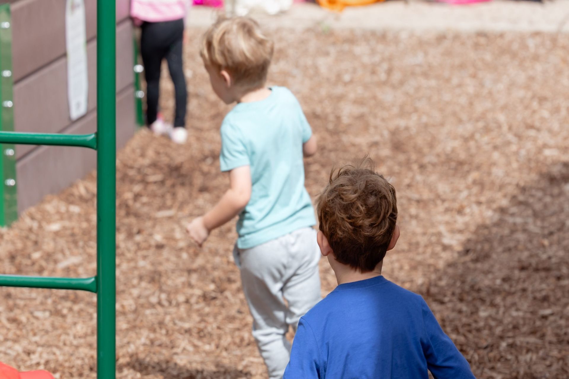 Children walking on a playground with wood chips, near green climbing bars and a slide.