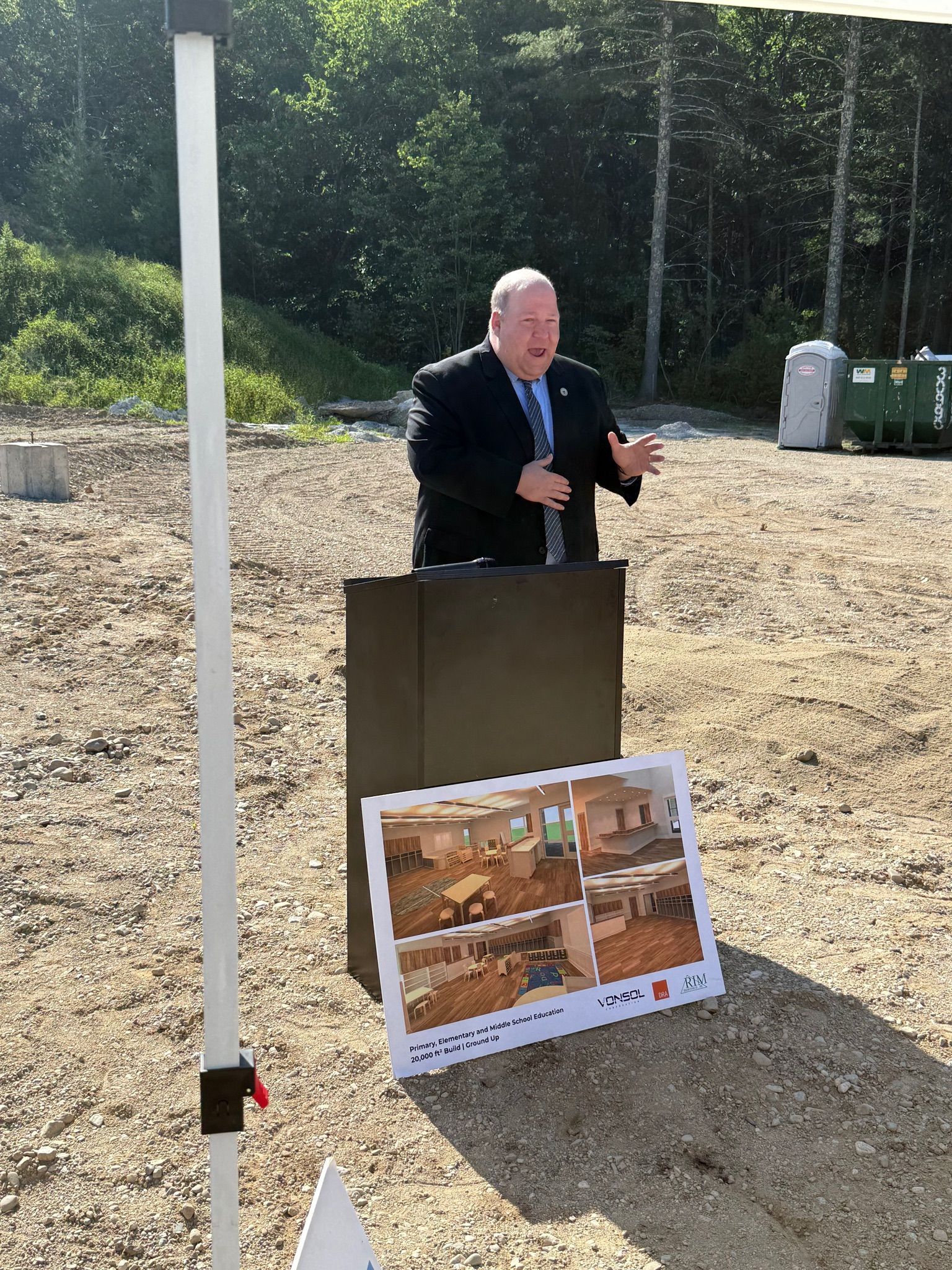 A man in a suit and tie is standing at a podium in a dirt field.