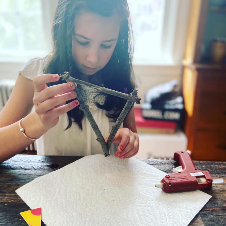 Montessori child crafting with twigs, focused, using a glue gun. Indoors, working on a tissue paper covered table.