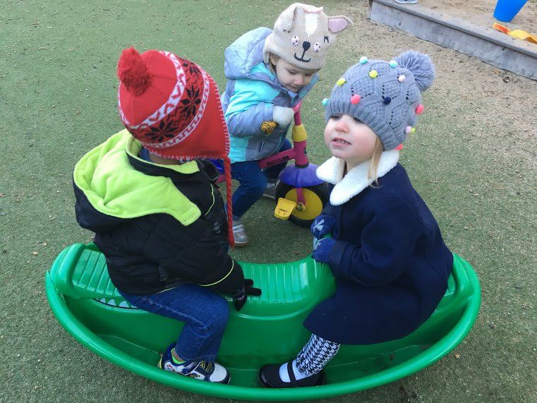 Three children in winter hats playing on a green playground rocking toy.