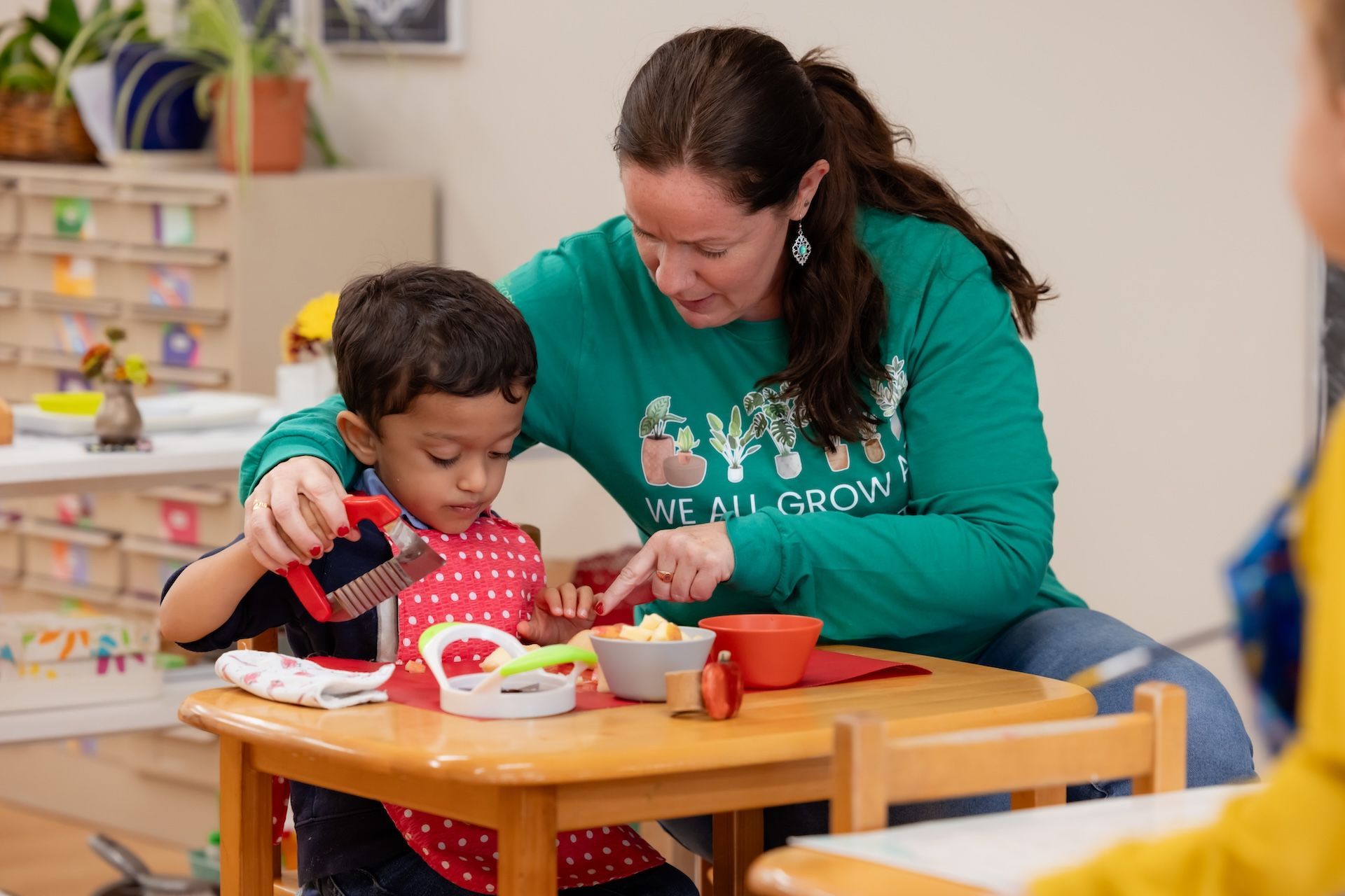 Montessori guide helps a child at a table with food and utensils. Both are focused on food preparation.