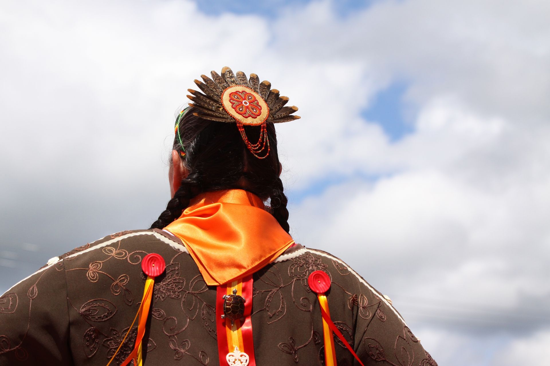 Person in traditional brown clothing and headdress, back to camera, under cloudy sky.