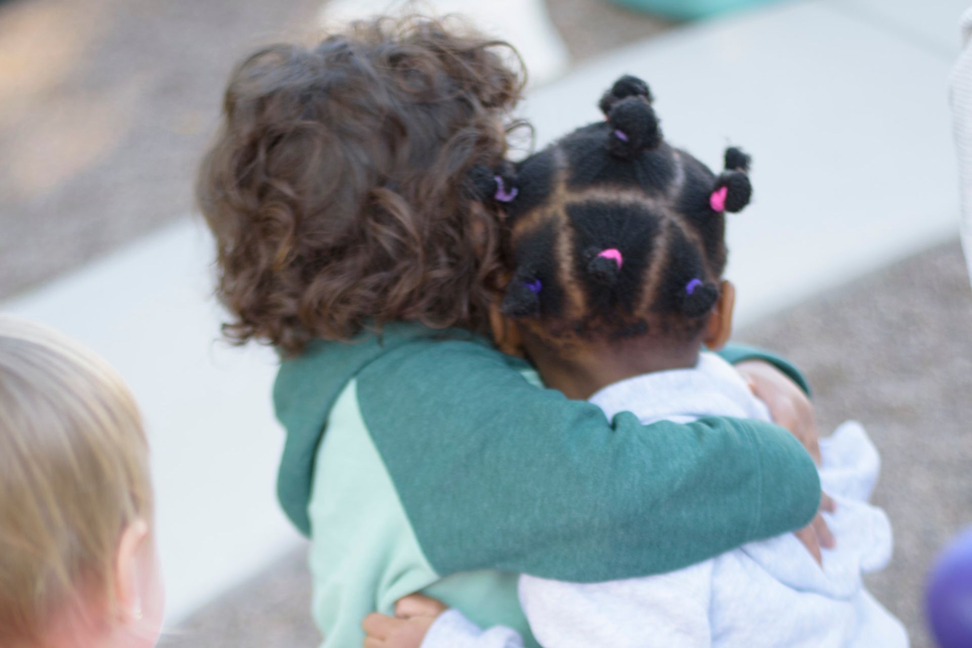 Two Montessori children hugging, one with curly brown hair, the other with braided hair and pink ties.