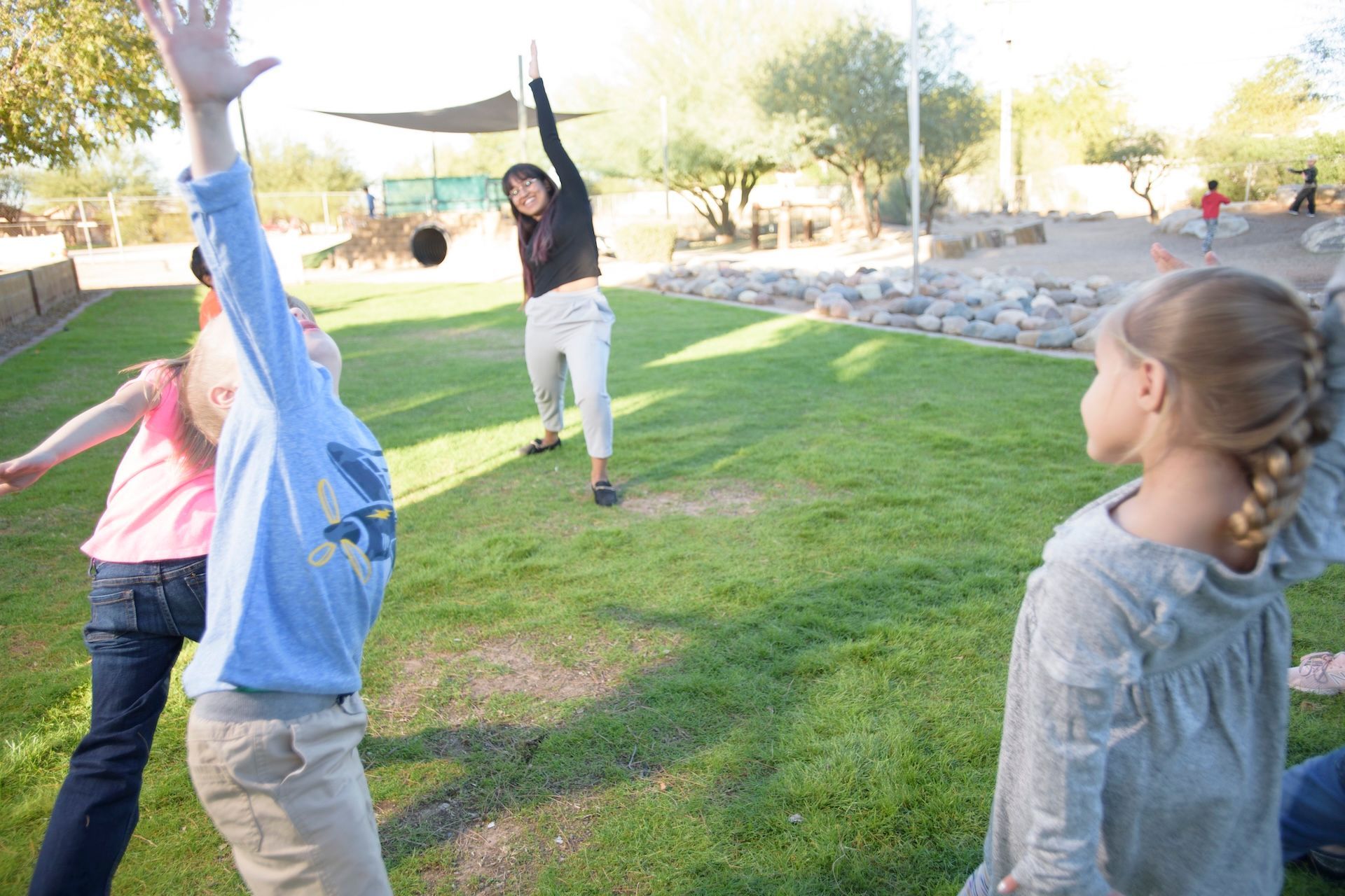 Montessori children playing a game on grass with a guide in a park. One child raises an arm.