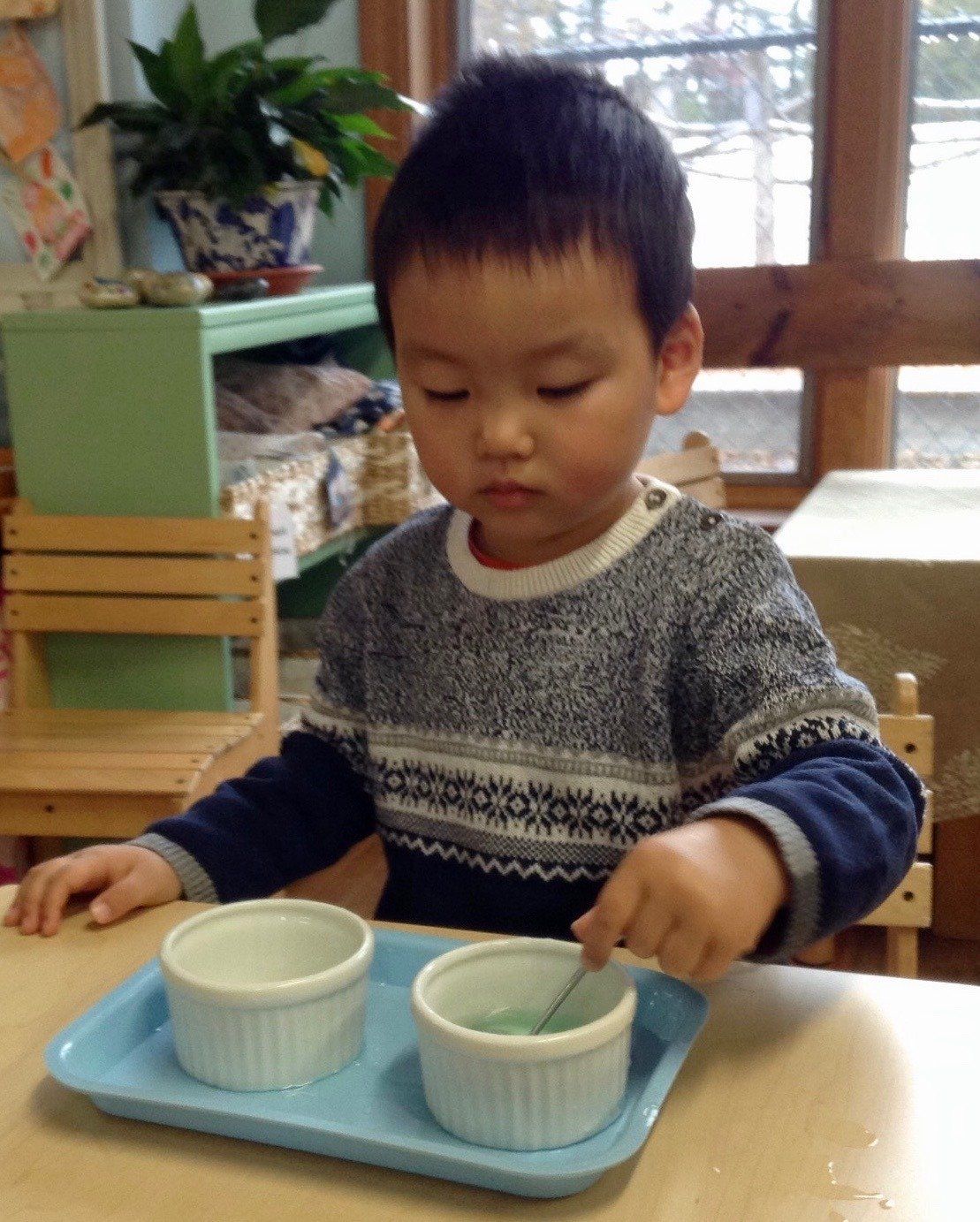 Montessori child stirring liquid in a bowl with a spoon on a blue tray.