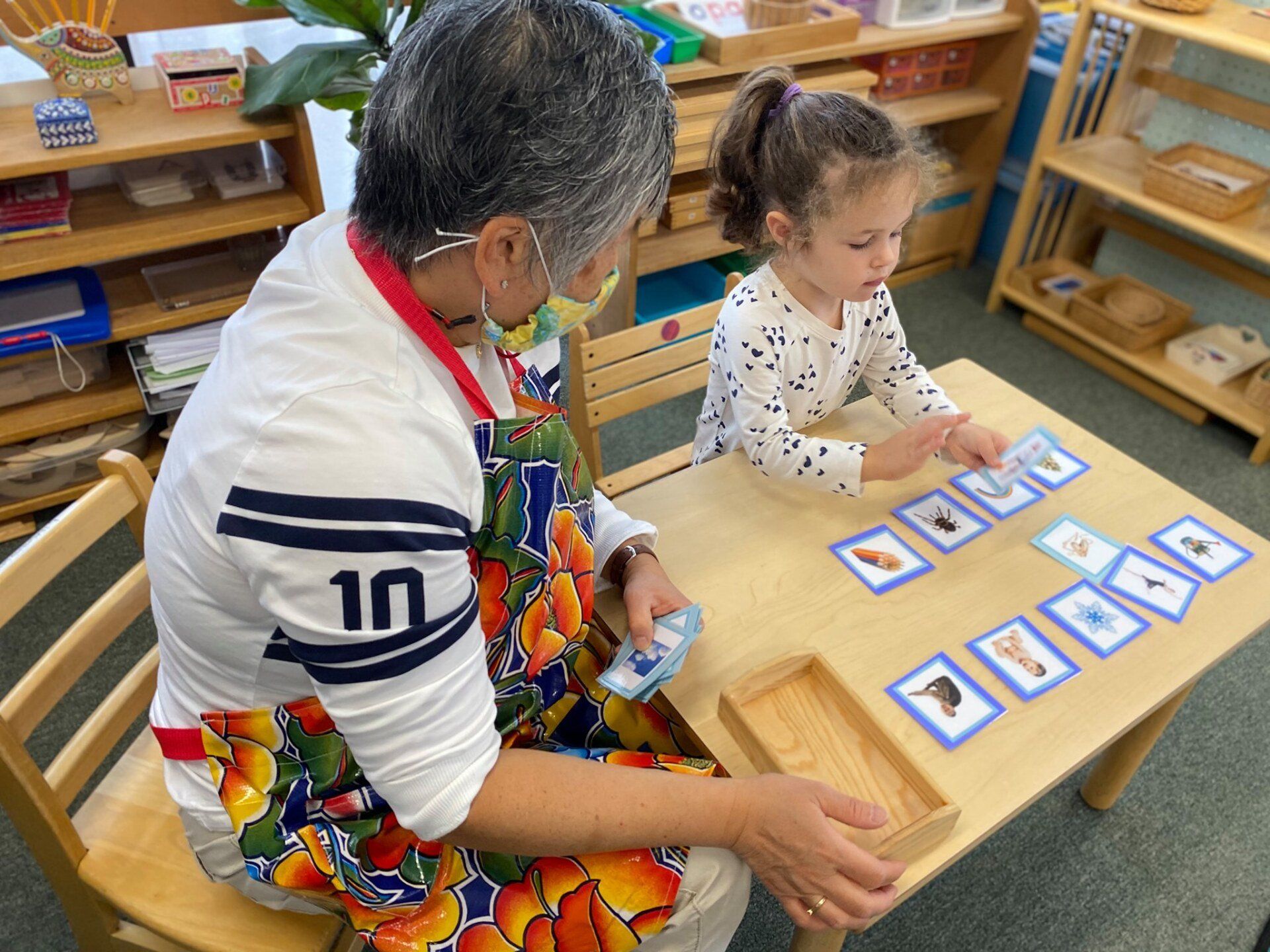 A montessori guide is sitting at a table with a little girl playing with montessori learning cards.