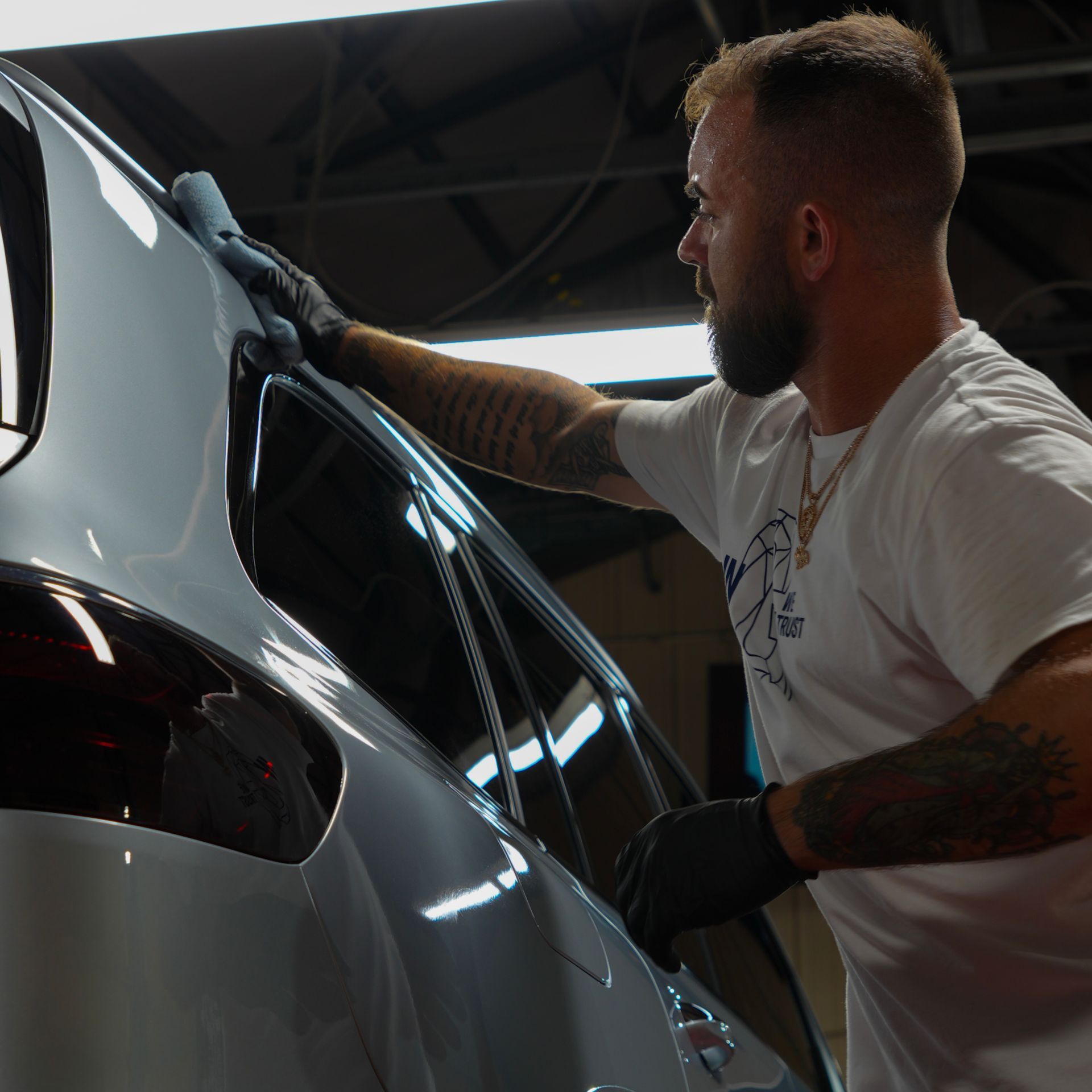 Detailer polishing a silver car in a dim garage, wearing gloves and a white shirt