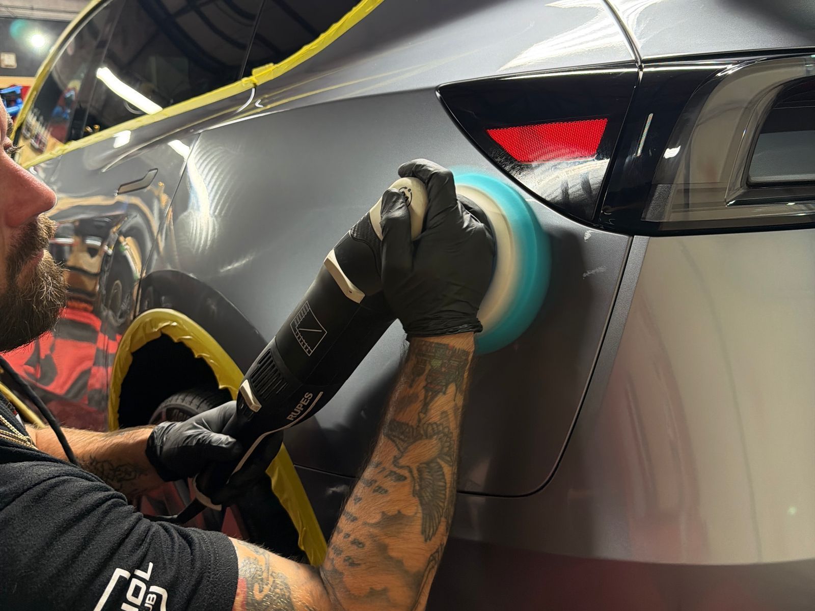 Detailer polishing a silver car’s rear panel with a buffer, yellow masking tape around the edge.