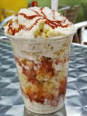Clear plastic cup of shaved ice topped with white cream and red syrup, with fruit and syrup layers inside.